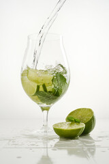 Clear shiny glass filled with with lime and mint and delicious Mojito cocktail composed on a white background. Pouring water into glass.  Shallow depth of field