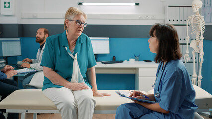 Senior woman having conversation with medical assistant at checkup appointment, female nurse taking...