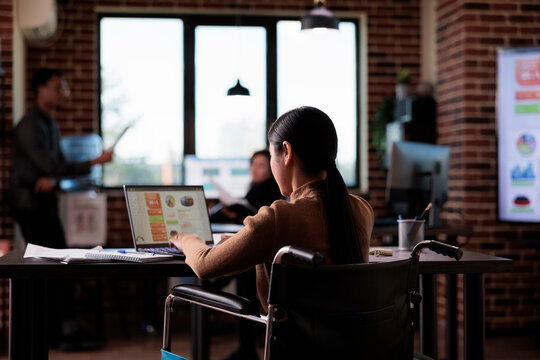 Female manager living with disability analyzing website on laptop, working with statistics in disability friendly office. Asian employee suffering from chronic impairment, using wheelchair.