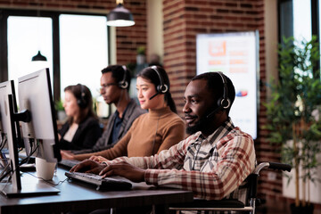 African american helpline operator with chronic impairment working at call center client care. Man wheelchair user giving telework assistance at customer service in disability friendly office.