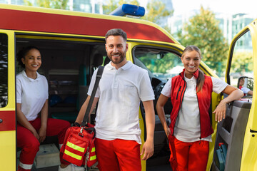 Three paramedics by the ambulance. Young woman and man smiling. Doctor is carrying a medical trauma...