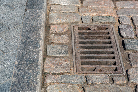rusted rectangular grating manhole drainage system on the pavement road nearby to the gray granite curb and stone tile pedestrian pavement close-up on dry weather, nobody.