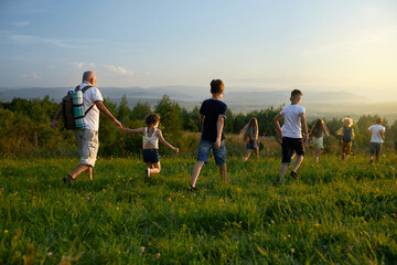Fototapeta premium Back view of kids with old man leading each other forward in hills. Male with gray hair wearing rucksack together with kids hiking, traveling, running ahead. Concep of active rest.