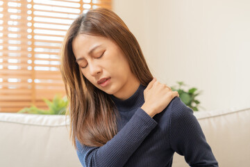 Body muscles stiff problem, asian young attractive woman, girl pain with back pain ache from work, holding massaging rubbing shoulder hurt or sore, painful sitting on sofa at home. Healthcare people.