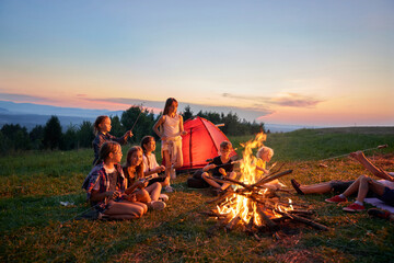 Side panoramic view of group of children sitting at campsite in mountains. Kids eating, having dinner, communicating, having rest near fireplace and rent. Concept of active rest. - obrazy, fototapety, plakaty