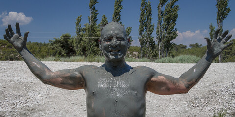 A man at a river mud bath, covered in mud, smiles and stretches his arms wide