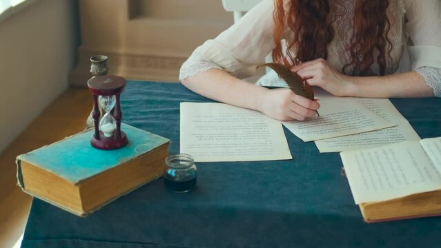 Medieval red-haired woman writer holds pen feather quill in hands close-up, face cropped, red lips. Girl sits at table writes letter on sheet paper. Vintage dress, old books. student studying in room
