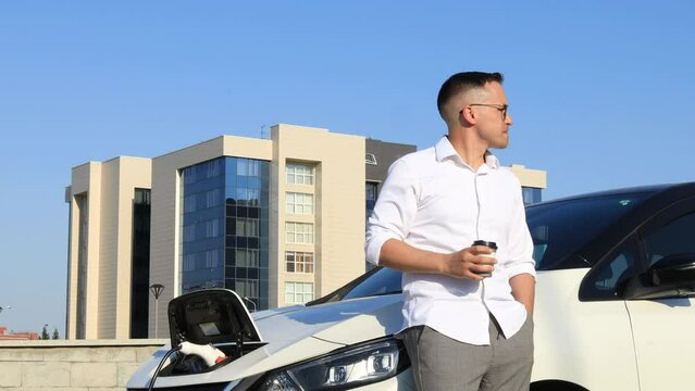 Young handsome man traveling in an electric car, stopping at a charging station, standing, plugging in a cable, browsing the internet on a smartphone, smiling joyfully while charing