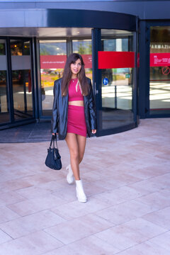 Portrait Of A Caucasian Girl In A Red Dress Walking Out Of The Revolving Door Of The Shopping Center, Fashion Portrait In The City