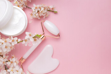 Container with bodycare and skincare cream, Gua sha stone, roller for face massage on a pink background with blooming cherry. Cosmetic facial skin care and spa. Natural treatment concept.