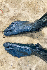 Pair of feet covered in black healing mud standing on sand. Top down view.