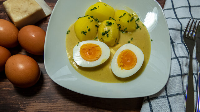 S&uuml;&szlig; saure Eier mit Petersilien Kartoffeln und So&szlig;e auf einem wei&szlig;en Teller auf Holztisch mit Tuch Besteck Speck unf rohen Eiern
