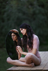 Yound brunette woman in bodysuit with mirror in hands sitting on wooden bridge