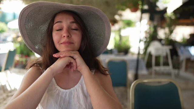 Young charming woman sitting on the left in sidewalk cafe talking in slow motion and looking at camera. Female Caucasian tourist in straw hat posing on sunny day on Cyprus outdoors
