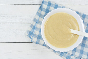 The first complementary food of child, baby nutrition. Oatmeal porridge for the baby (multi-grain) from ground cereals in white plate on blue cloth on a wooden background. Space for text.