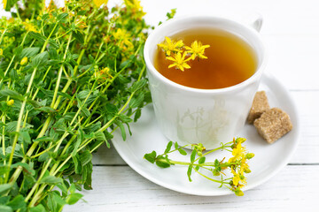 A drink (infusion, decoction) of St. John's wort in a white cup on a saucer on a white wooden background close-up. Herb and flowers of the medicinal plant Hypericum, Hypericaceae.