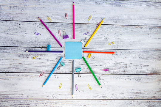 Bright, multicolored pencils for drawing in an album, colored paper clips, a blue notebook for notes and clips are folded neatly in a circle on a light, gray and wooden background.