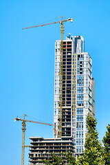White and blue glass skyscraper under construction with yellow tower crane along it and smaller concrete base of the building and crane next to it against clear blue sky with warm light of the sun