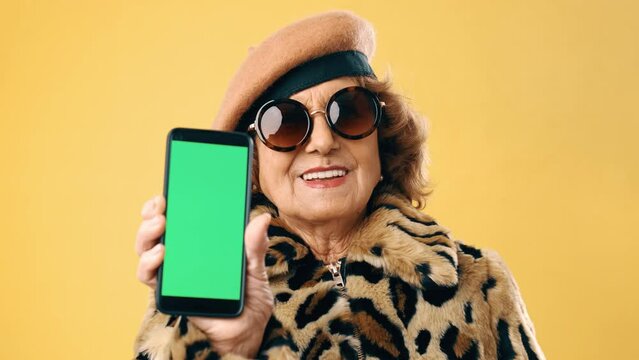 Stylish senior woman smiling while showing a mobile phone with green screen over an isolated background.