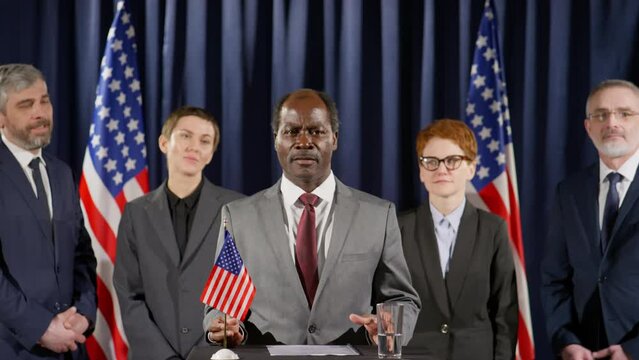 African American politician standing at rostrum with US flag on it and giving speech as his team applauding during press conference