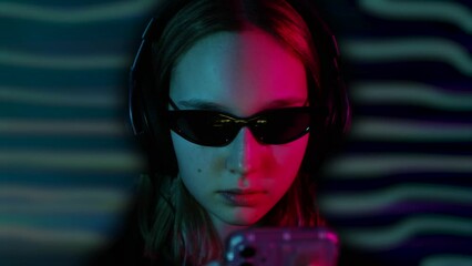 Close-up face of girl in headphones and black sunglasses with mobile phone against multicolored neon waves background. Teenage girl listen music and use phone