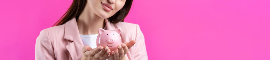 A cute girl in a pink jacket is holding a 3d piggy bank in her hands. Beautiful close-up portrait in studio on pink.