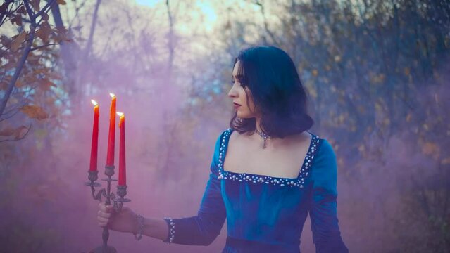 creative art video shooting. Portrait fantasy woman queen in dark autumn forest, holding burning candlestick in hands, red candles. Background forest autumn trees filled with fog, pink smoke checkers