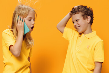 cute, cheerful children, brother and sister, of school age in yellow clothes stand on an orange background and the girl is enthusiastically talking on the phone.