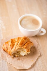 Bitten croissant and cup of cappuccino coffee on wooden table. Breakfast or coffee break in cafe