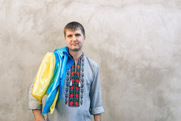 Obraz na płótnie Canvas Portrait of a man holding the blue and yellow national flag of Ukraine on his shoulder and looking away. The guy in a traditional embroidered shirt on a background of a concrete wall. Copy space.
