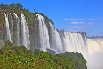 Naklejka premium Foz do Iguaçu Falls in Brazil. Bulky water continuously falling. Fog in the air and vision blurred by fog. International tourism, UN preservation area. Clear blue sky and green vegetation at the bott