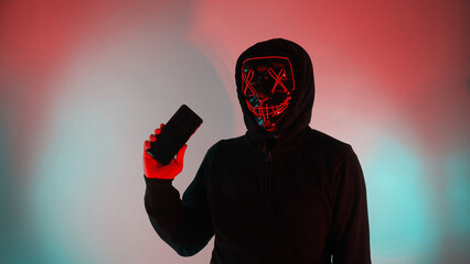 Anonymous hacker and face mask with smartphone in hand. Man in black hood shirt trying to hack...