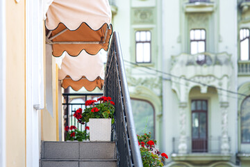 facade of building with an external staircase to balcony with black railing and decor on wall and decorative sun visors above windows and decorative flower pots with blooming red flower, nobody.