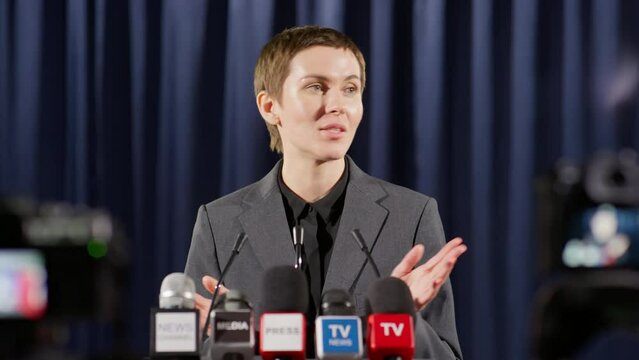 Female politician standing at rostrum and talking in mics while answering questions of TV journalists in front of cameras during press conference