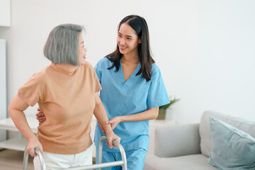 Young asian female caregiver helping senior woman to walk with walker at home.