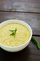 Raw risoni pasta in a white bowl with a sprig of rosemary on a wooden table. The concept of Italian cuisine. Risoni is suitable for salads and soups. Vertical orientation.