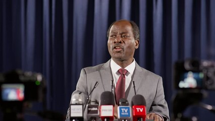 African American politician in formal suit standing at podium with mics and holding speech in front of TV cameras during press conference