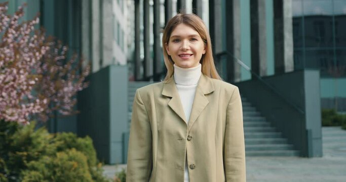 Portrait of caucasian woman looking at camera while standing outside office building. Businesswoman beautiful stylish dressed in jacket posing and smiling near office centre.