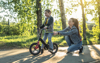 Fototapeta na wymiar Mom helps a child boy learn to ride a two-wheeled bicycle in the park. A pleasant children's summer sports vacation