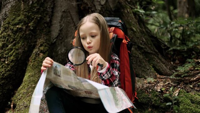 Cute little independent girl with a backpack in a forest studies a tourist map with a magnifying glass in search of a hiking route. 