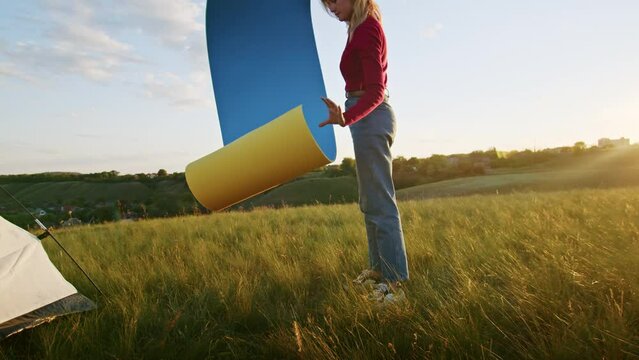 a young woman happily set up a tent on the mountain hills during sunset, for rest after a hike. a woman lays out a karimat. rest of one person in nature, in the mountains.