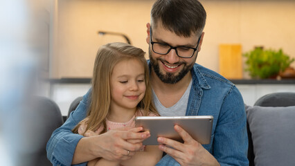 Father and little girl use digital tablet sitting at home. Computer games, surfing internet, social media, buy in online store