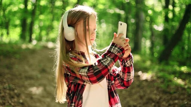 Cute little girl hold smartphone, watching social media and taking photo in the park. Smiling child texting on her phone. 