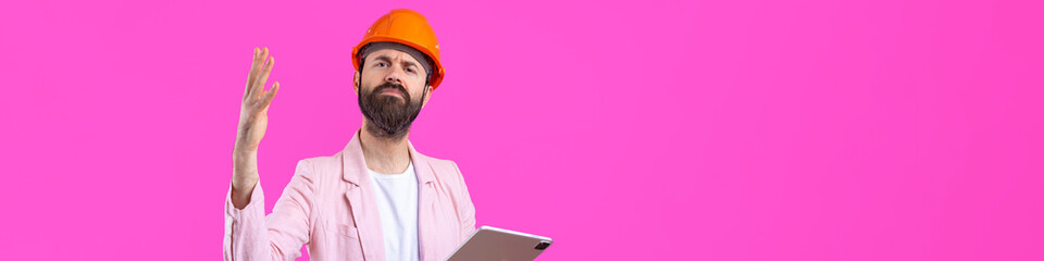 Portrait of young construction engineer wear orange hard hat, in a pink jacket standing on red studio background. A man with a tablet in his hands.