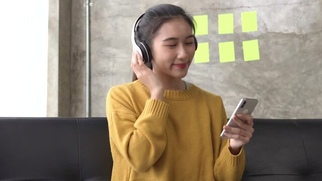 Happy asian woman relaxing and using overhead headphones to listen to music from smart phone sitting on sofa.