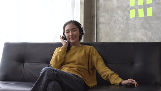 Happy asian woman relaxing and using overhead headphones to listen to music from smart phone sitting on sofa.