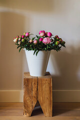 Big fresh pink and white peony blooming bouquet in a white metal bucket on a wooden table in a light bright modern beautiful apartment. Spring home decor or present idea. Vivid summer colors.