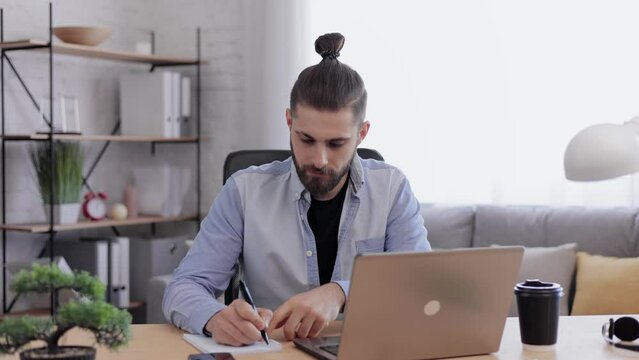 Handsome bearded man studying online using laptop web camera, writing lecture in notebook, talking with teacher. Remote working,home education.Online meeting,video call,video conference,courses online