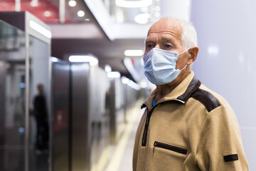 Fototapeta na wymiar Elderly man in protective medical mask is standing on platform and waiting train in underground