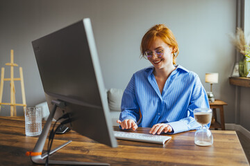 Smiling young female employee busy working on computer typing writing business letter to client,...